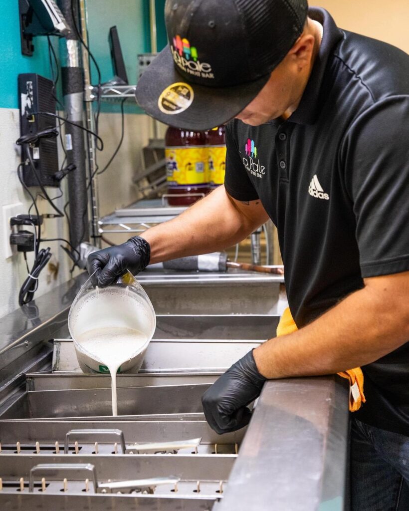 A worker wearing gloves pours liquid mixture into metal molds used to make frozen paletas.