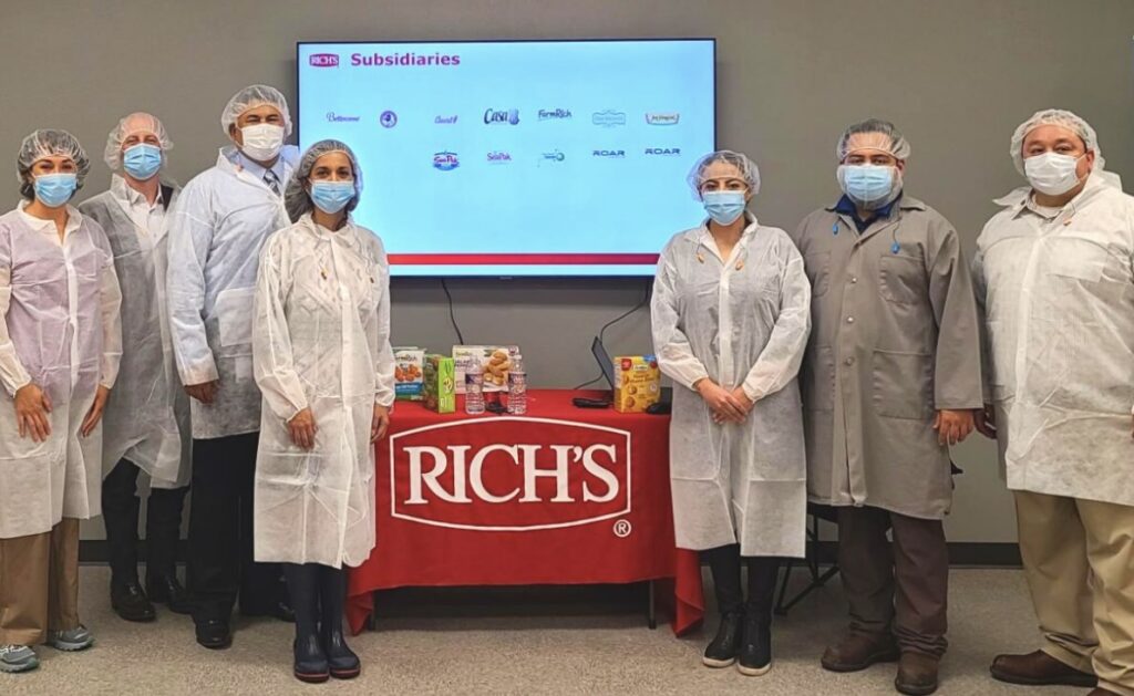 Un grupo de responsables de desarrollo económico y representantes del sector manufacturero, que llevan redecillas para el pelo, mascarillas y batas protectoras, se encuentran en una sala de reuniones junto a un expositor con marcas de productos, en lo que representa una reunión celebrada en 2021 relacionada con la ampliación prevista de una planta de producción alimentaria de Brownsville.