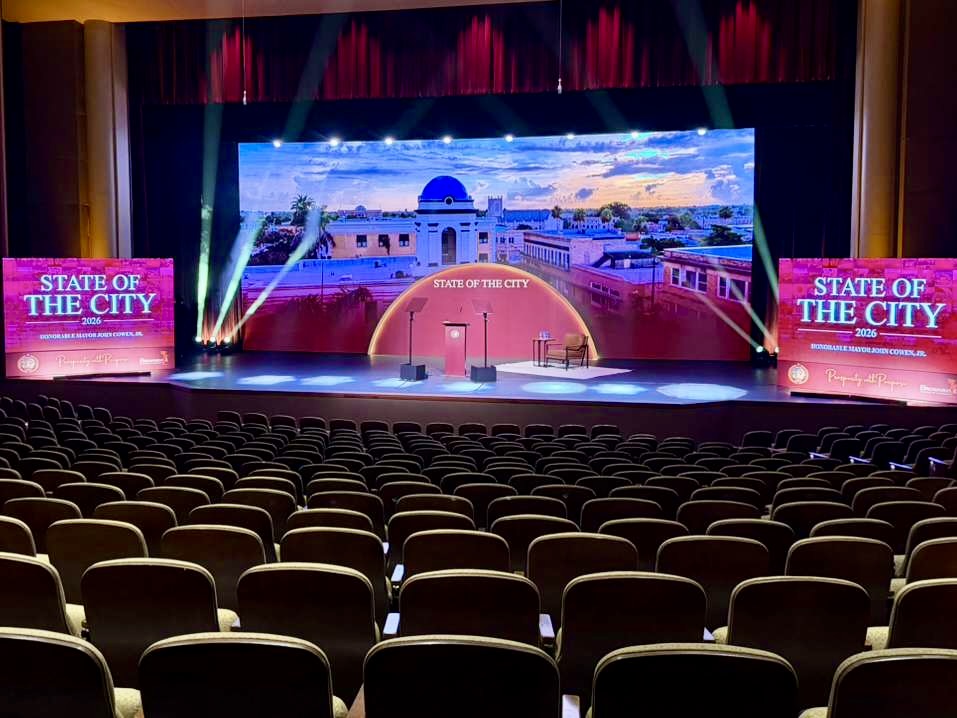 A theater stage displays “State of the City 2026” with lighting, screens, and a city backdrop, viewed from rows of empty seats.