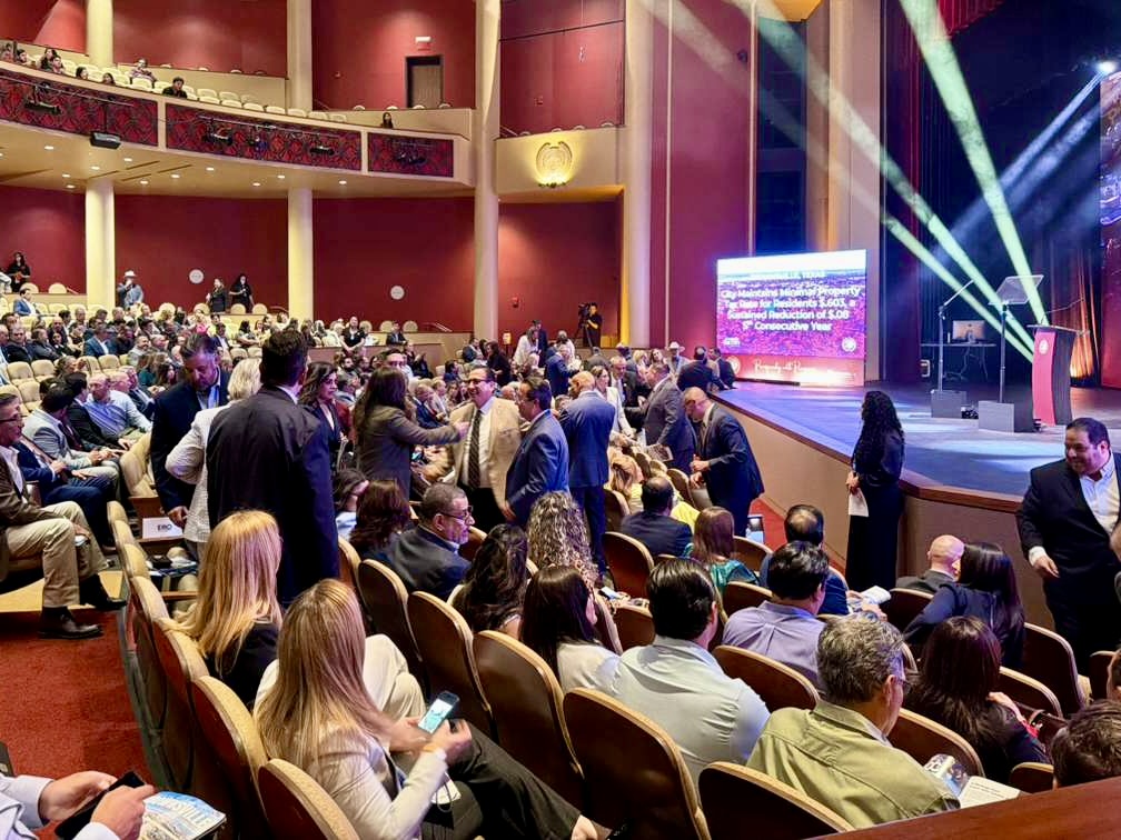 Audience members fill a theater-style auditorium, mingling and taking their seats before a formal city event.