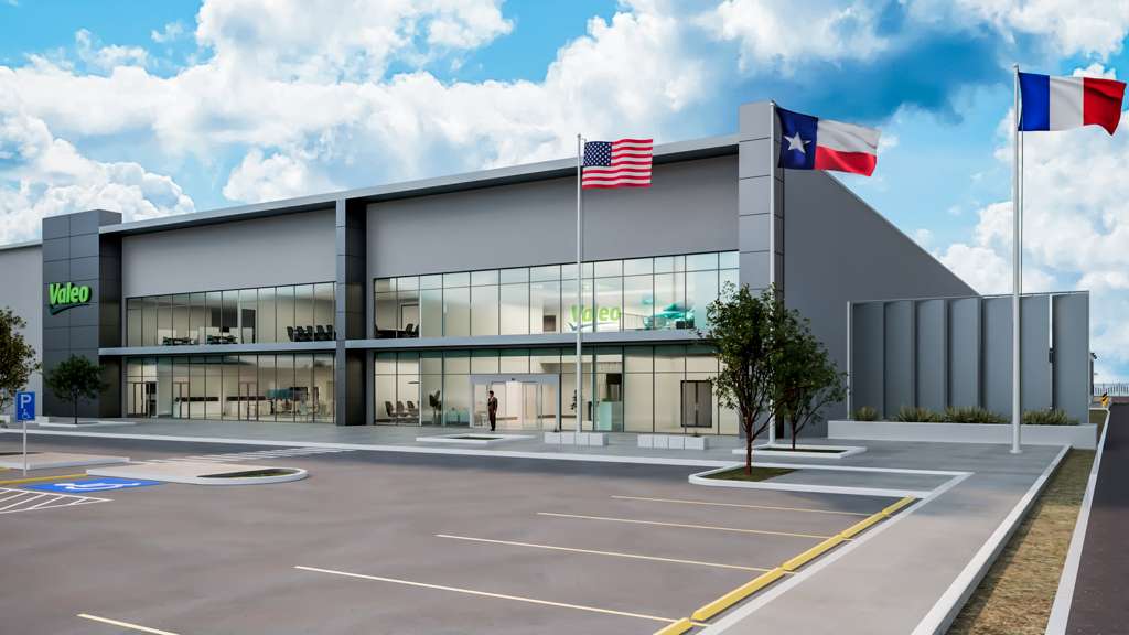 Exterior rendering of a modern industrial manufacturing facility with large glass windows, a gray facade, and flags of the United States, Texas, and France displayed outside, representing Valeo’s planned plant in McAllen.