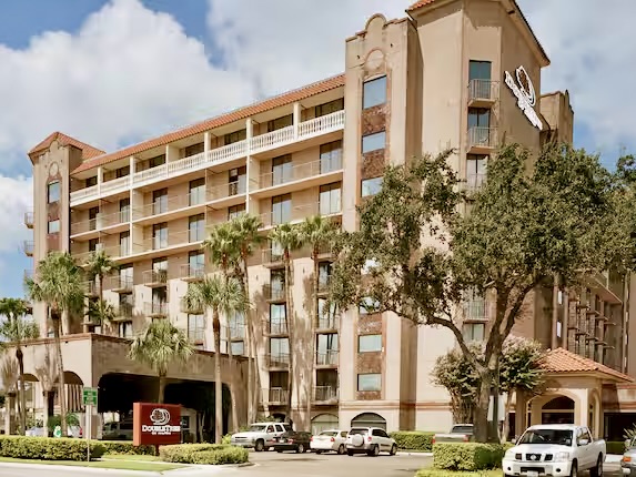 Vista exterior del hotel DoubleTree by Hilton de McAllen, en la que se aprecia un edificio de varias plantas con palmeras, una entrada cubierta y vehículos aparcados en la parte delantera.