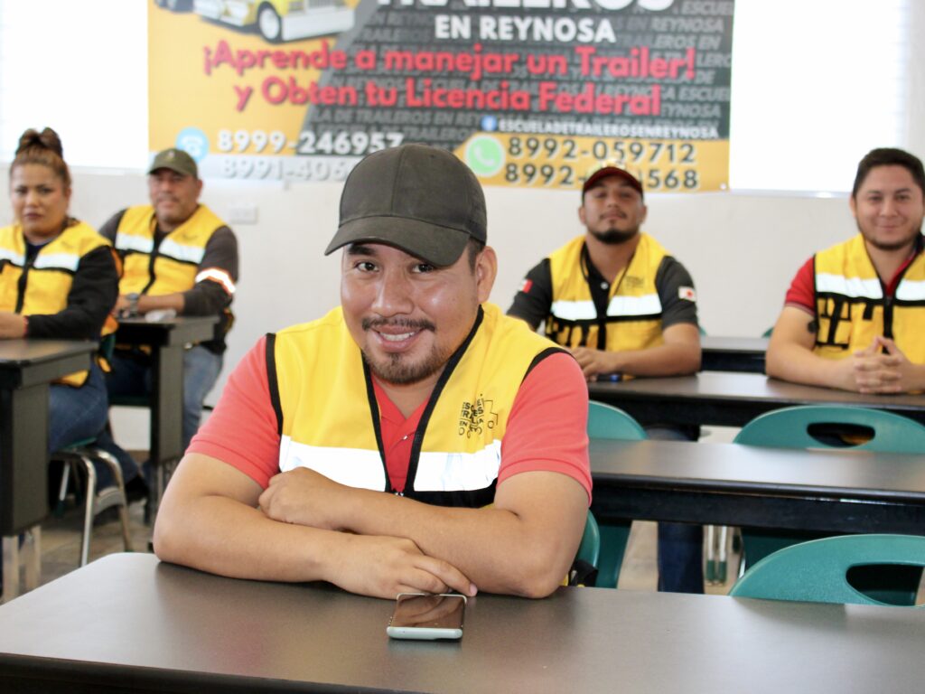 Un alumno con un chaleco de seguridad amarillo está sentado en un pupitre del aula, sonriendo a la cámara, mientras otros alumnos se sientan detrás de él.