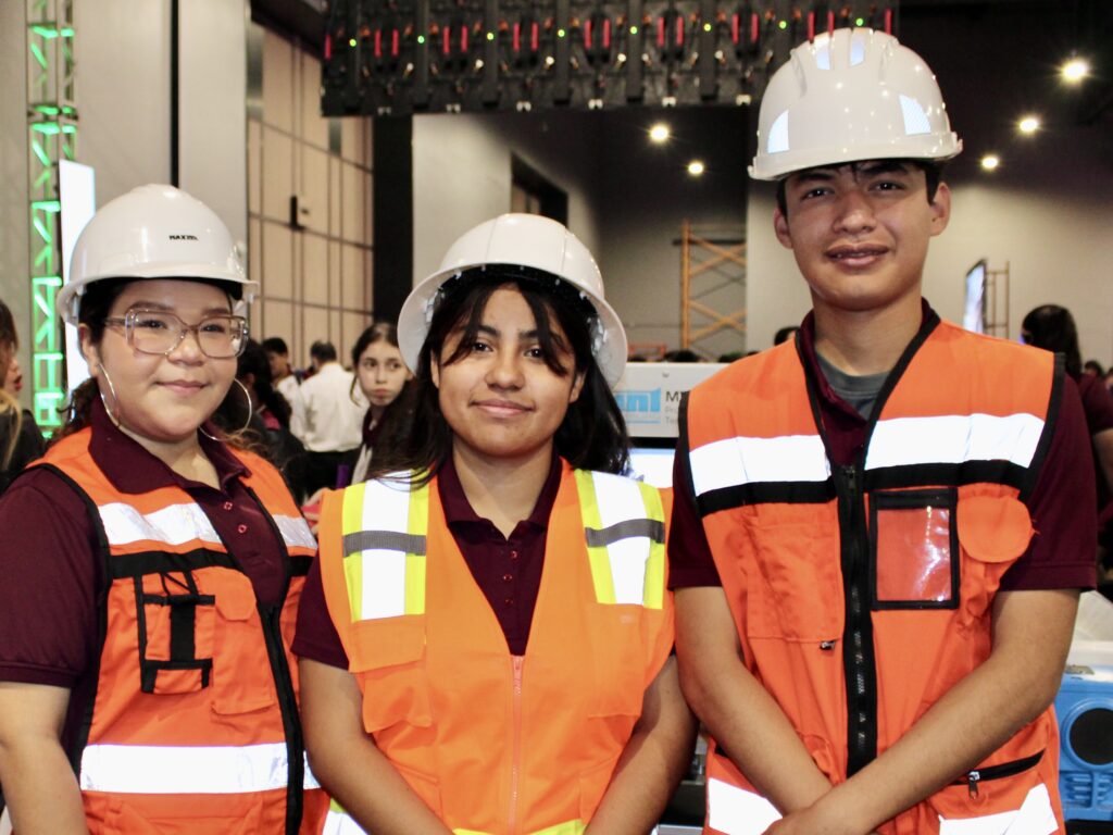 Three students wearing hard hats and safety vests pose together at an education expo.