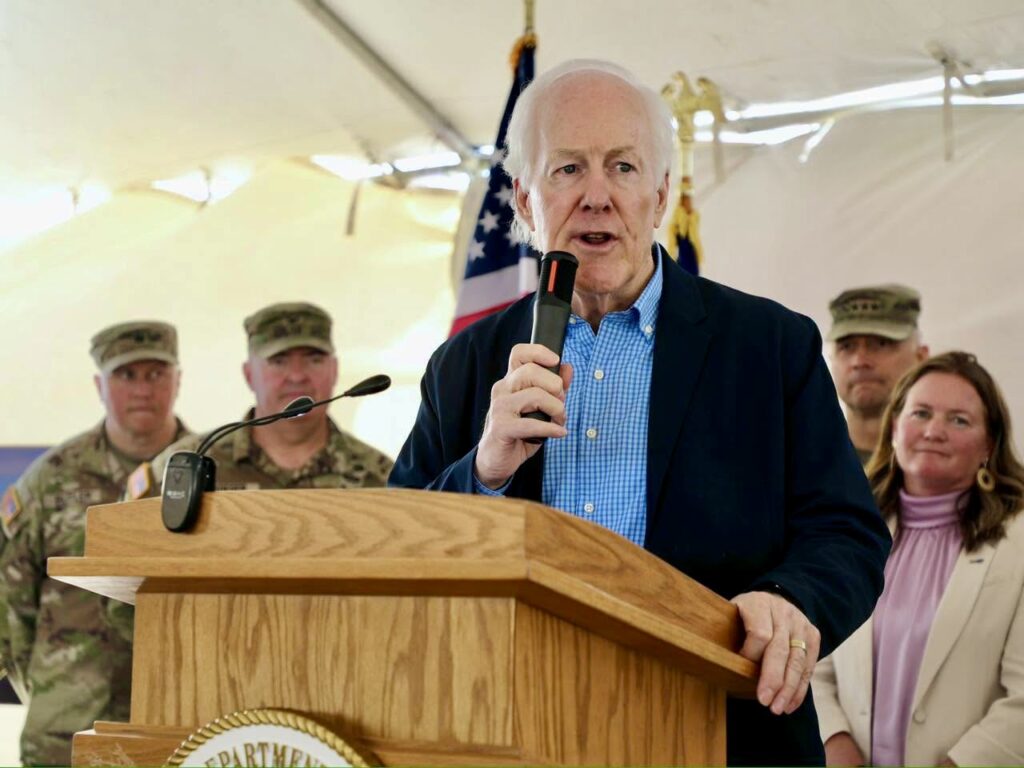 El senador John Cornyn habla desde un atril durante un acto al aire libre, con personal militar y autoridades de pie detrás de él.