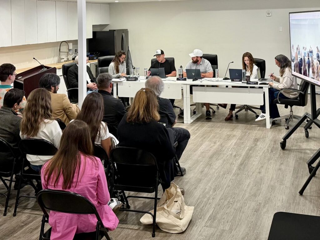 Los responsables municipales se sientan a una mesa durante una reunión de la Comisión Municipal de Starbase, mientras los asistentes observan desde sus sillas en una pequeña sala de reuniones.