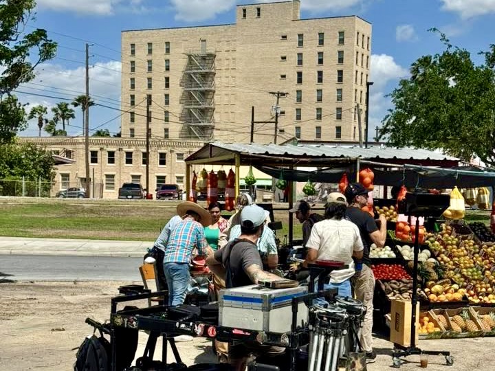 Film crew members stand around cameras and production equipment at an outdoor market-style set with fruits and vegetables in the Rio Grande Valley.