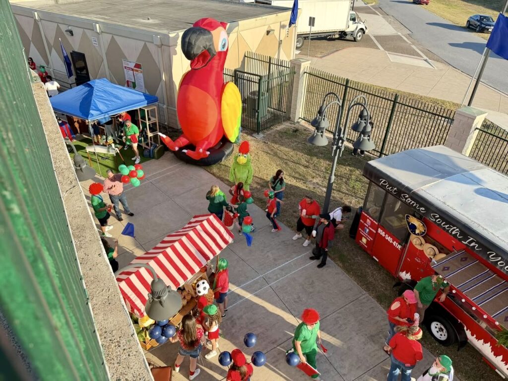 Vista aérea de un grupo de personas reunidas alrededor de una instalación festiva al aire libre en Harlingen durante una sesión fotográfica para la campaña del Mundial, con decoraciones coloridas, puestos y un gran loro hinchable.