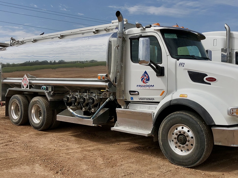 Un gran camión cisterna blanco para combustible, con el letrero «Freedom Fuels», está aparcado en un terreno de tierra, dejando a la vista el lateral del depósito y el equipo de transporte.