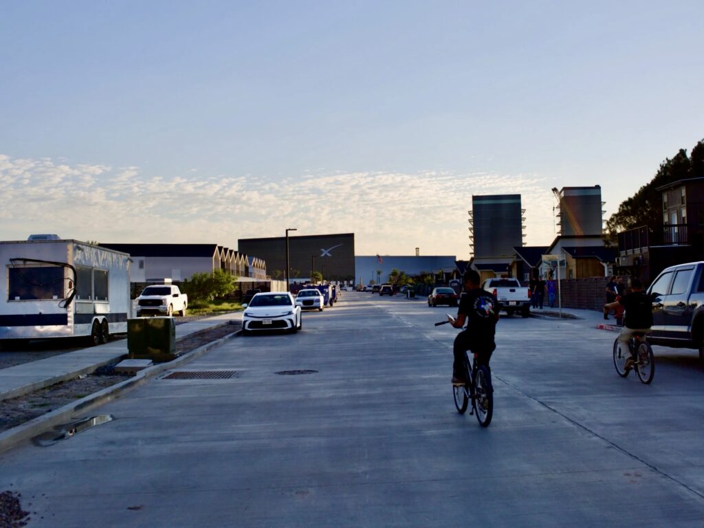 Una calle residencial de la ciudad de Starbase, cerca de la playa de Boca Chica, muestra viviendas, vehículos aparcados y ciclistas, con las instalaciones de SpaceX visibles al fondo.