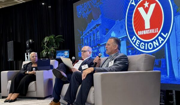 Three panelists sit on a stage during a conference in Brownsville, with America First Refining COO Tom Ramsey speaking into a microphone. A large screen behind them displays a “YTexas Regional” logo.