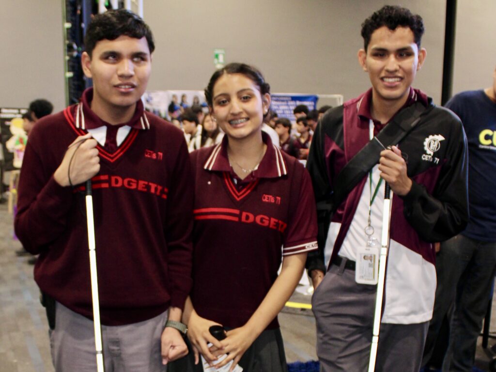 Three high school students stand together at an education expo, two holding white canes indicating visual impairment.