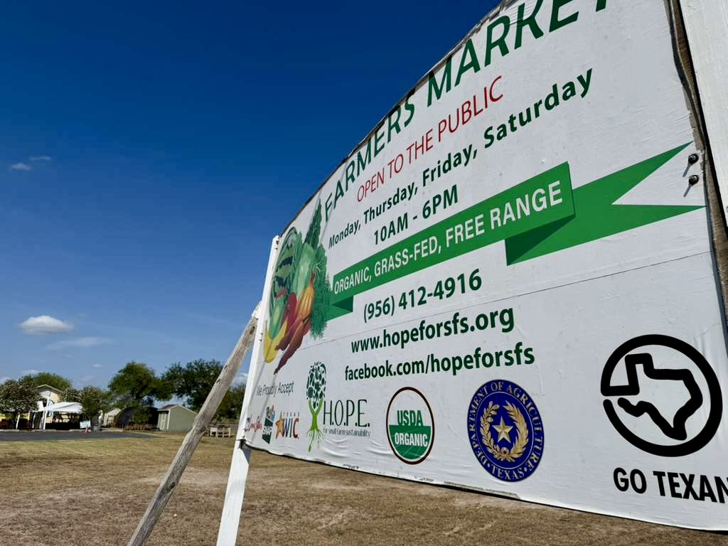 Close-up of a farmers market sign in Harlingen listing hours, contact information, and agricultural program affiliations.
