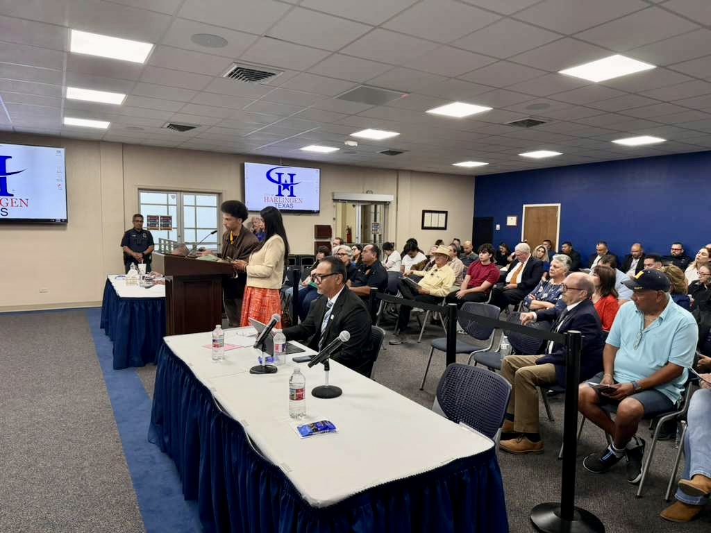 A man speaks at a podium during a Harlingen city meeting while residents and officials listen in a packed room.