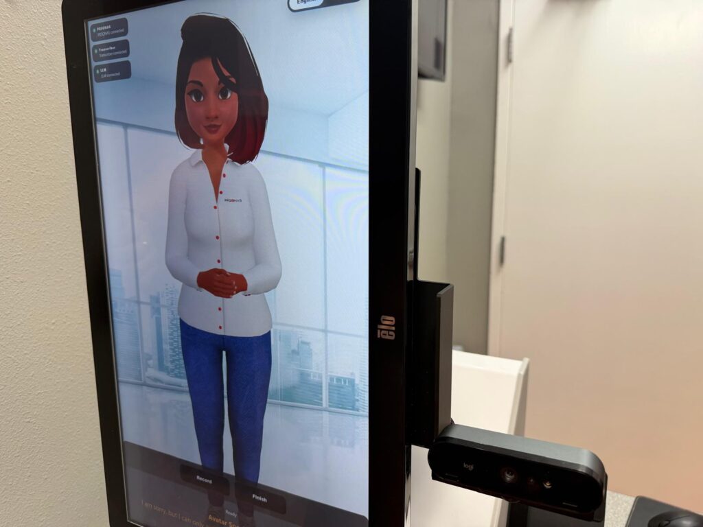 Primer plano de un avatar generado por IA que aparece en una pantalla digital con un sensor de cámara, utilizado para el registro de pacientes en una clínica médica.