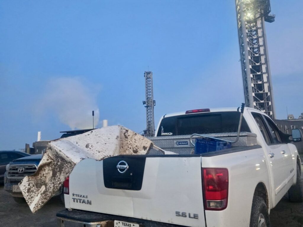 Large piece of debris resting on the back of a white pickup truck near Starbase launch infrastructure.