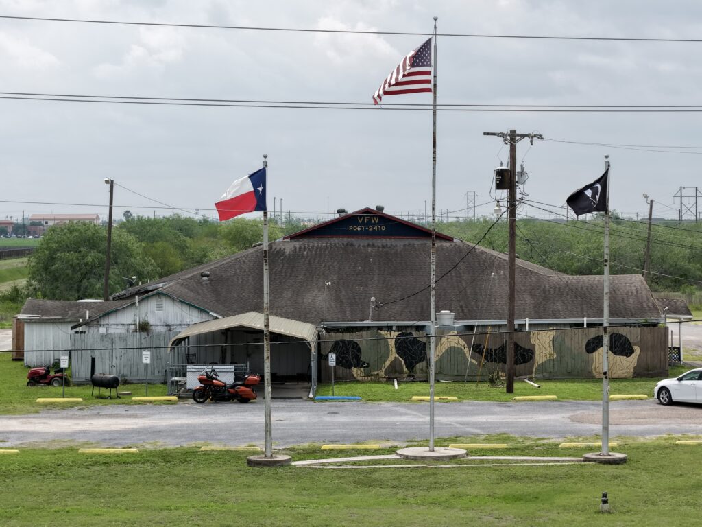 Exterior view of the aging VFW Post 2410 building in Harlingen with U.S., Texas, and POW/MIA flags flying in front of the deteriorating structure.