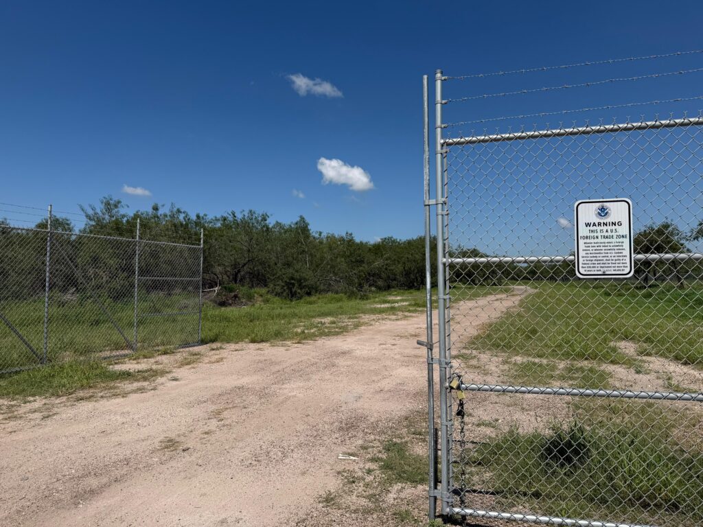 A gated dirt-road entrance to a Foreign Trade Zone property surrounded by brush land and chain-link fencing in north McAllen.