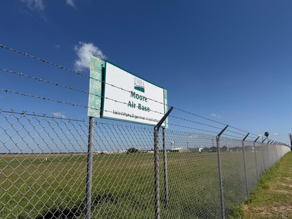 A USDA Moore Air Base sign stands behind a chain-link fence near open land in north McAllen under a bright blue sky.