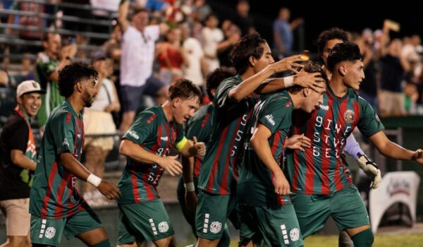 RGV Red Crowns soccer players in green and red uniforms celebrate on the field as fans cheer from the stands during a nighttime match at Harlingen Field.