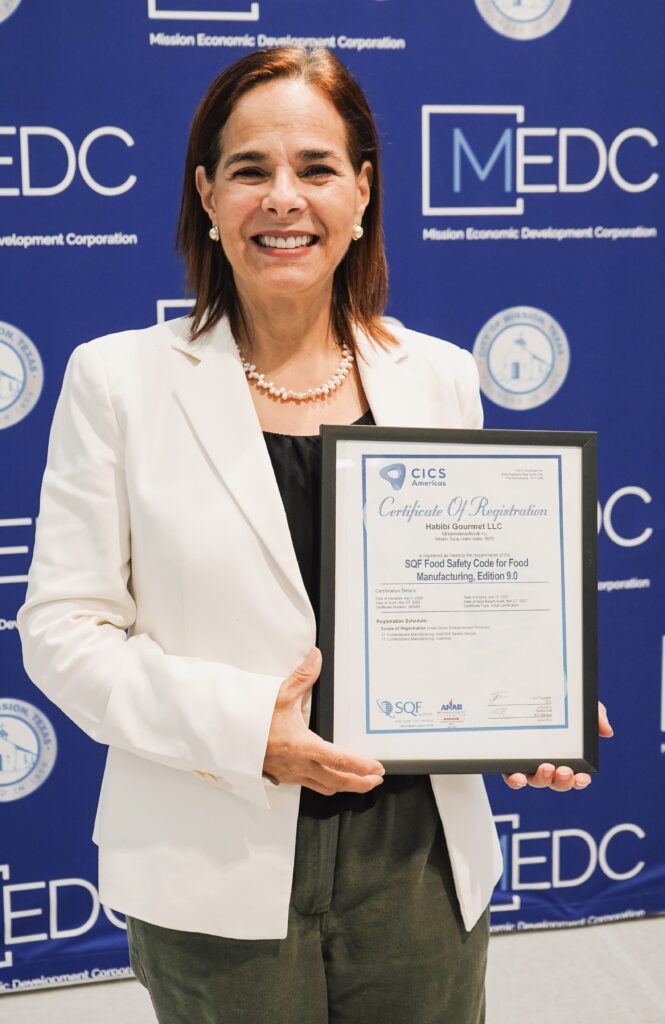 Pilar Gonzalez holds Habibi Gourmet’s Safe Quality Food certification certificate in front of a Mission Economic Development Corporation backdrop.