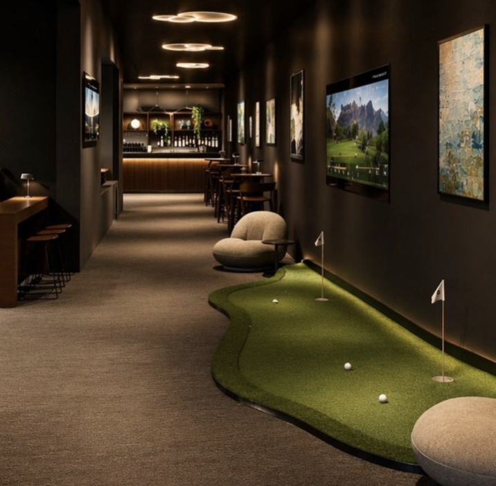 Una representación de un salón de golf cubierto que cuenta con un putting green, zonas de descanso, televisores y un bar.