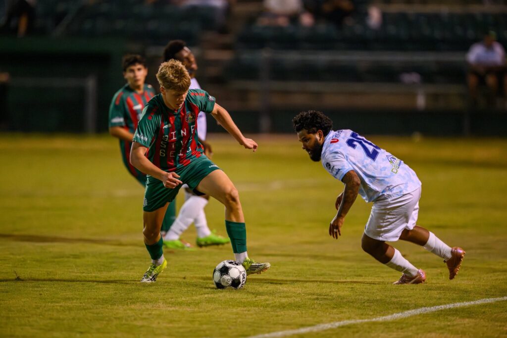 An RGV Red Crowns player in a green-and-red striped uniform dribbles past a defender during a nighttime soccer match at Harlingen Field.