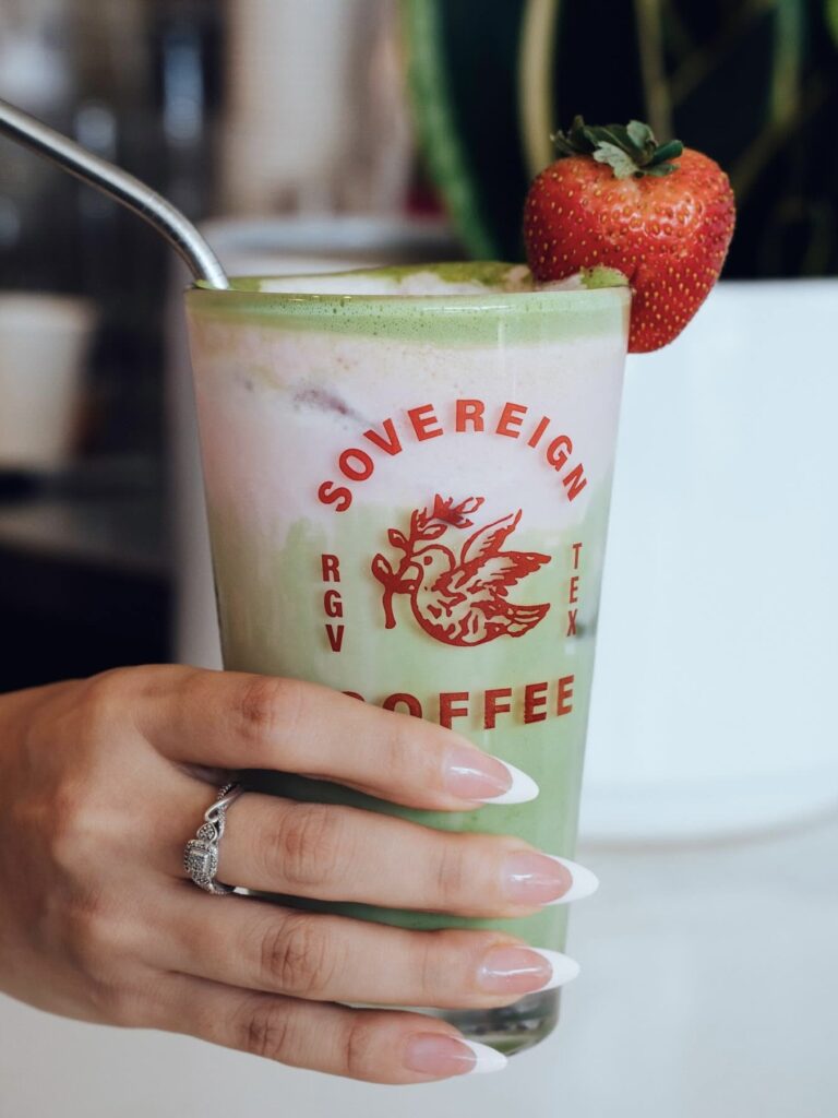 A hand holds a green Sovereign Coffee drink garnished with a strawberry.