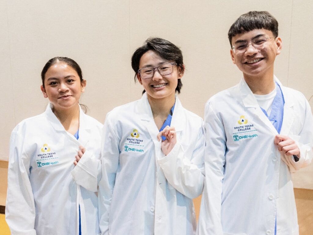 Three South Texas College nursing students wearing white coats with South Texas College and DHR Health logos.