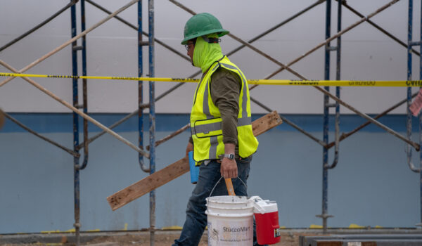 A construction worker wearing a hard hat and safety vest carries lumber and supplies at a job site.