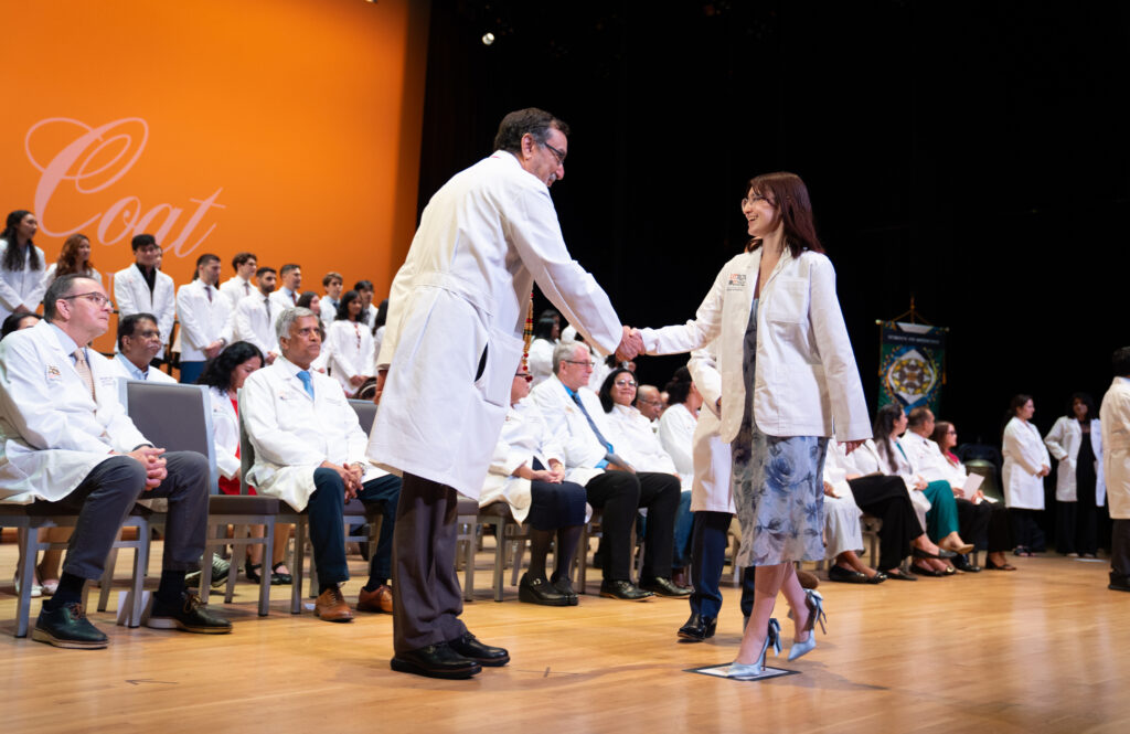 A faculty member shakes hands with an incoming UTRGV School of Medicine student after presenting her white coat during the 2026 White Coat Ceremony. Faculty, students and guests look on from the stage at the UTRGV Performing Arts Complex in Edinburg.