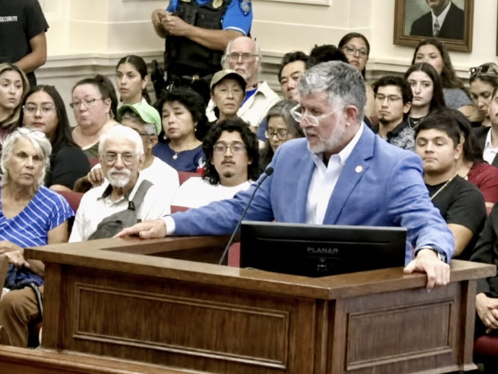 Rafael Vela, wearing a blue jacket, addresses Brownsville city commissioners from a podium in a crowded meeting room.