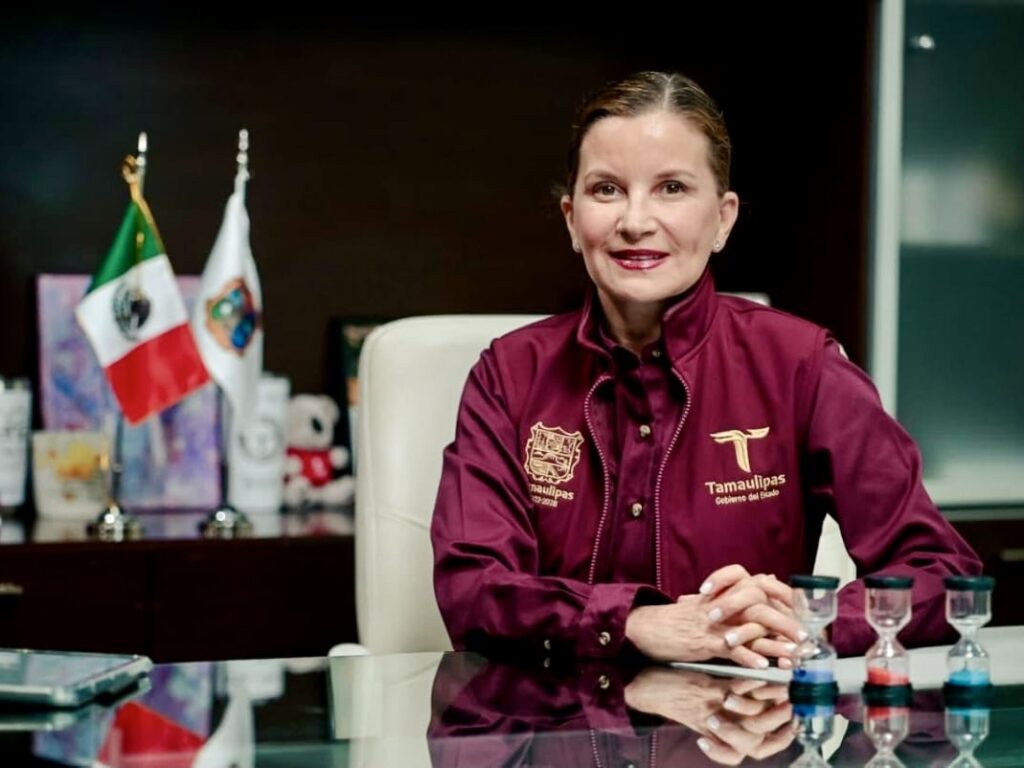 La secretaria de Economía de Tamaulipas, Ninfa Cantú Deándar, está sentada frente a un escritorio con las banderas de México y de Tamaulipas detrás de ella.