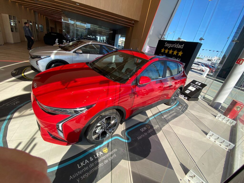 A red vehicle and a silver sedan are displayed inside the KIA Reynosa showroom.