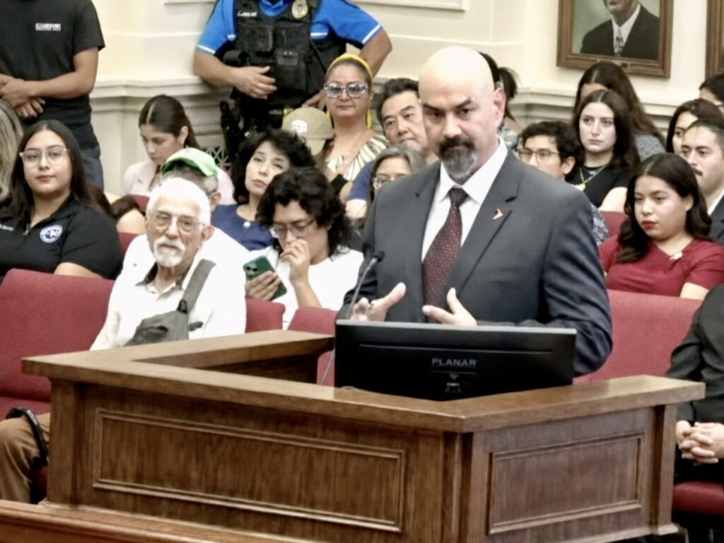 Gilberto Salinas, wearing a dark suit and tie, speaks at a podium during a crowded Brownsville City Commission meeting.