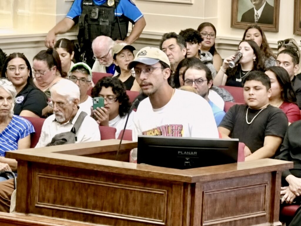 Alexis Diaz, wearing a baseball cap and white shirt, speaks at a podium during a Brownsville City Commission meeting.