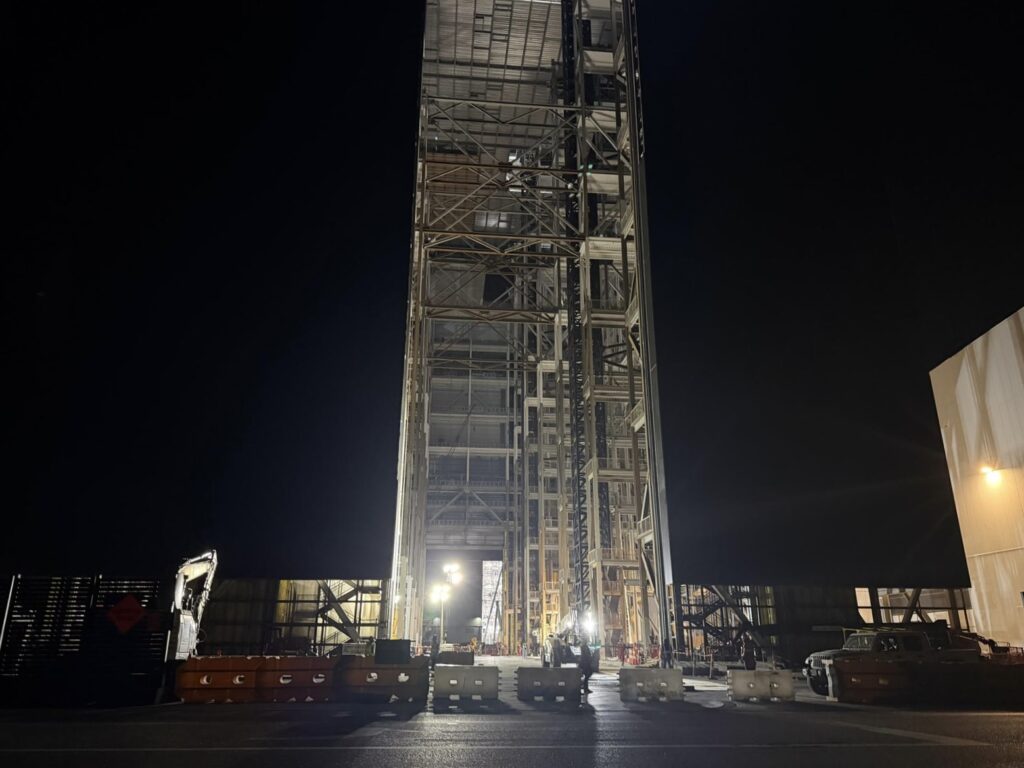 A tall SpaceX structure under construction at night, illuminated by work lights with construction equipment and barriers below.